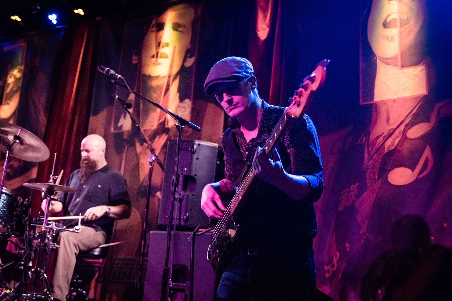 Musicians perform on stage with artistic portrait banners in the background.