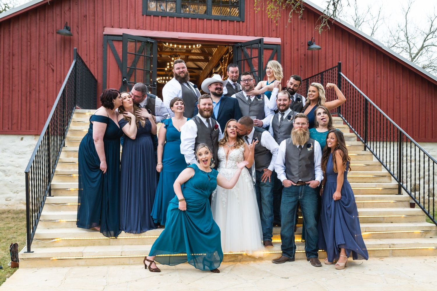 Large wedding party poses joyfully on rustic barn steps with bride in white and bridesmaids in blue.