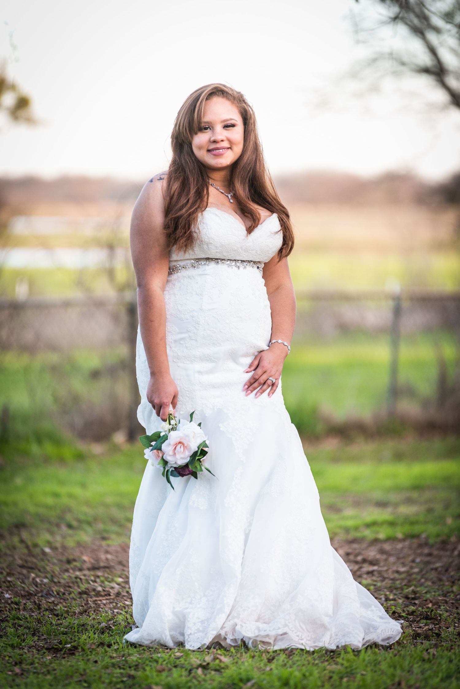 Bridal portrait in white lace gown holding pink roses against natural outdoor backdrop.