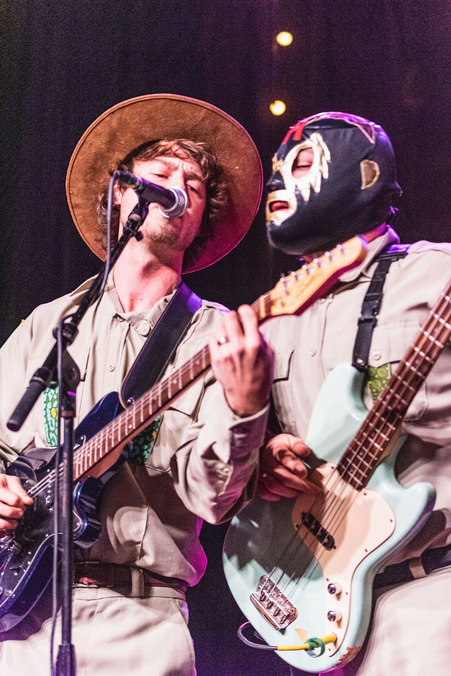 Two musicians perform on stage, one in cowboy hat and one in mask, playing guitars under stage lights.