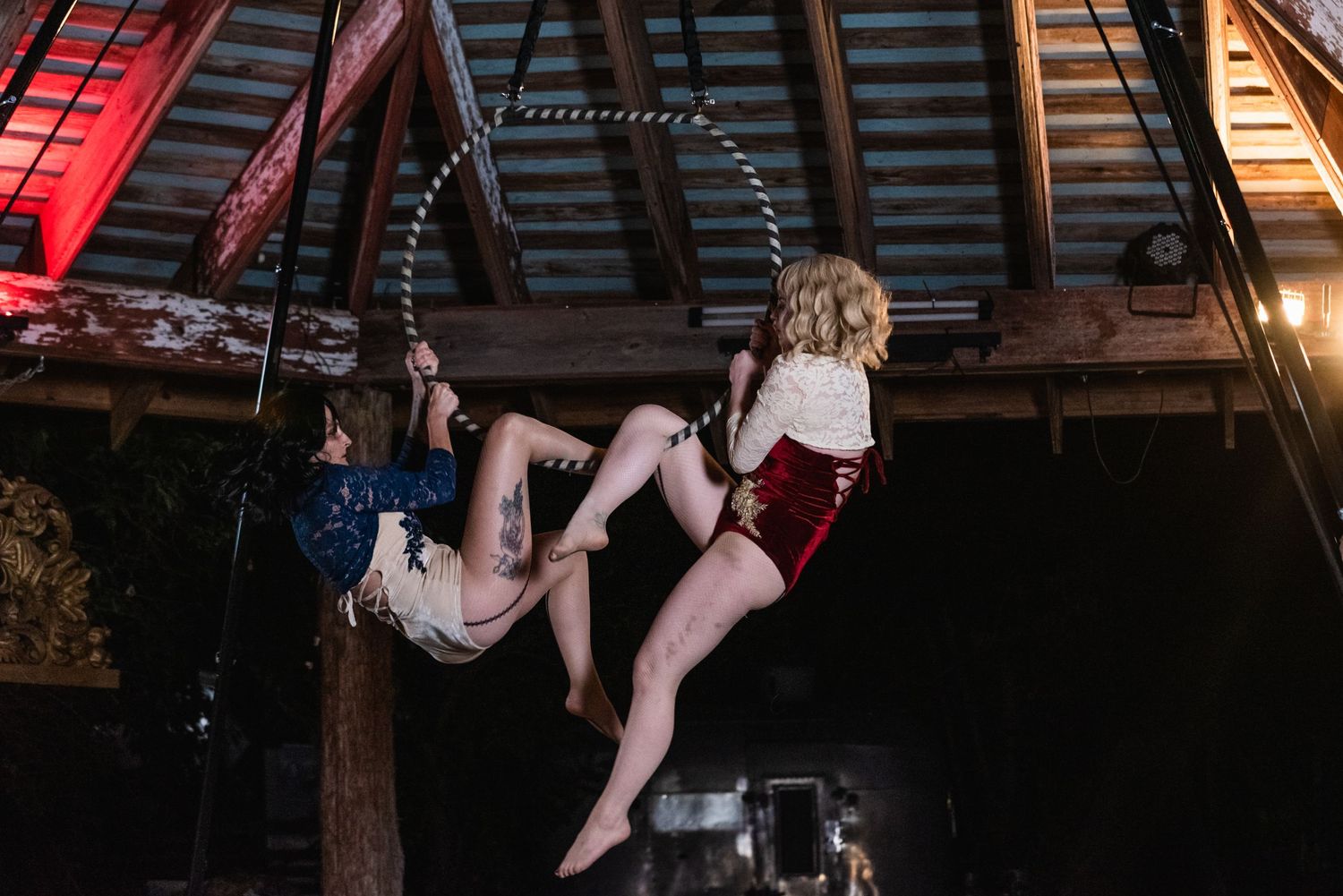 Two aerial performers practice acrobatic moves suspended from ropes in rustic venue.
