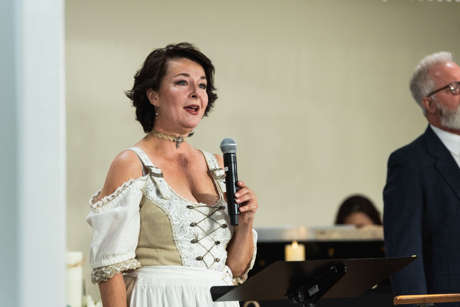 A performer in a white renaissance-style dress sings into a microphone during a show.