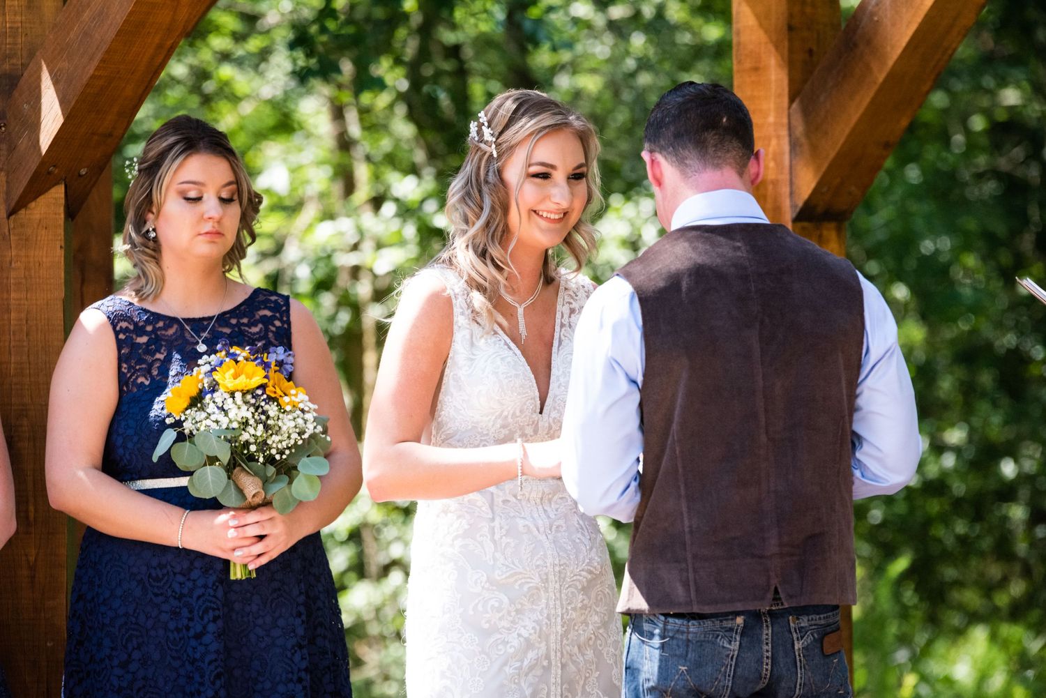 Beautiful outdoor wedding ceremony under wooden pergola with navy-dressed bridesmaid.