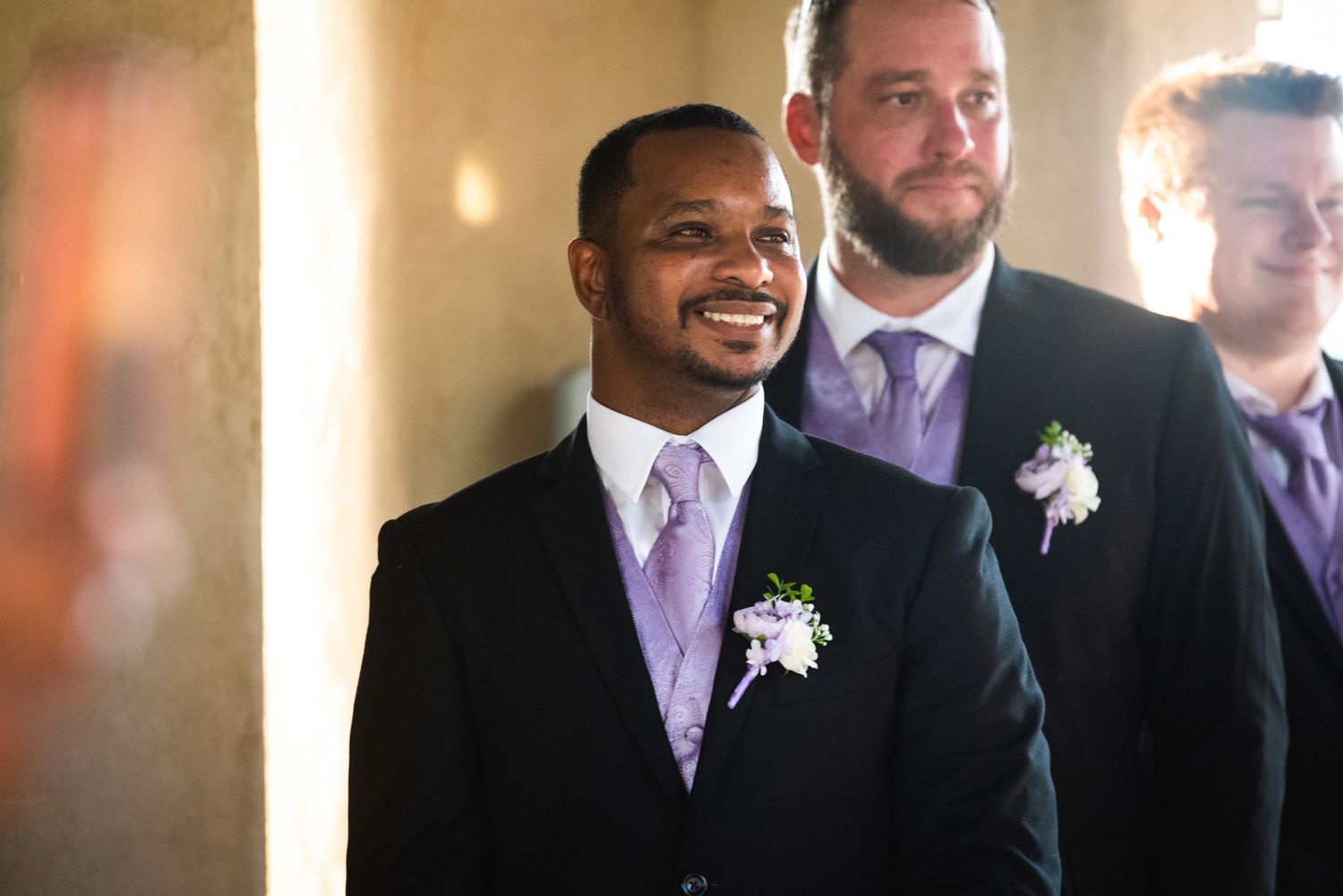 Formal wedding party members smile while wearing light purple ties and white boutonnieres indoors.