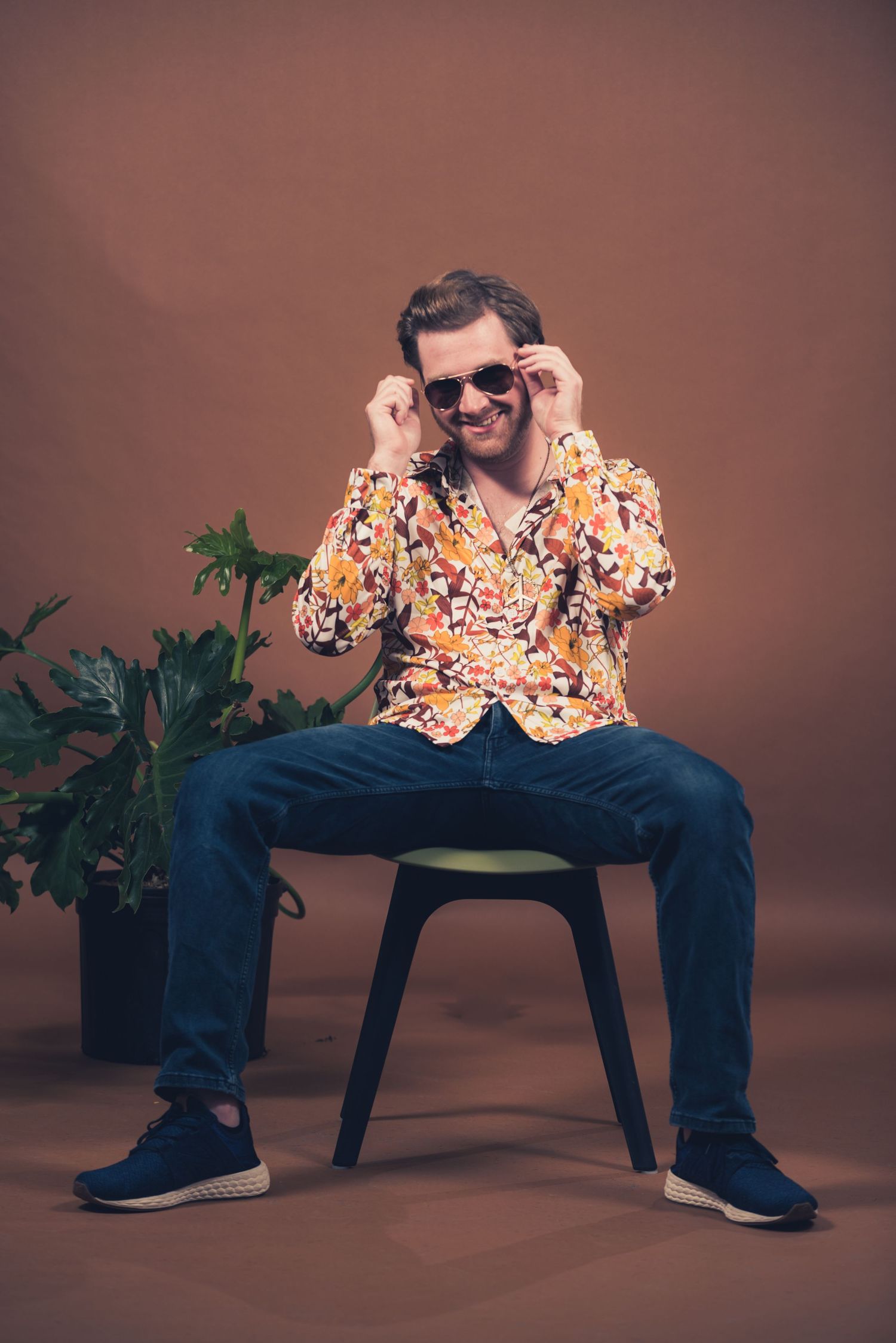 Person in floral shirt adjusts sunglasses while sitting on chair with plant in background.