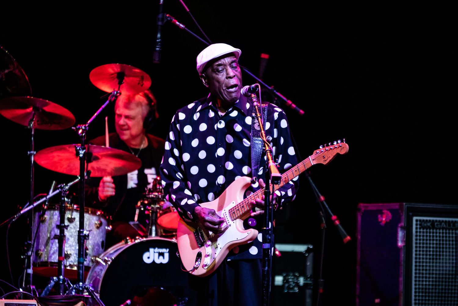 Rock musician in polka dot shirt performs with band under red stage lighting.