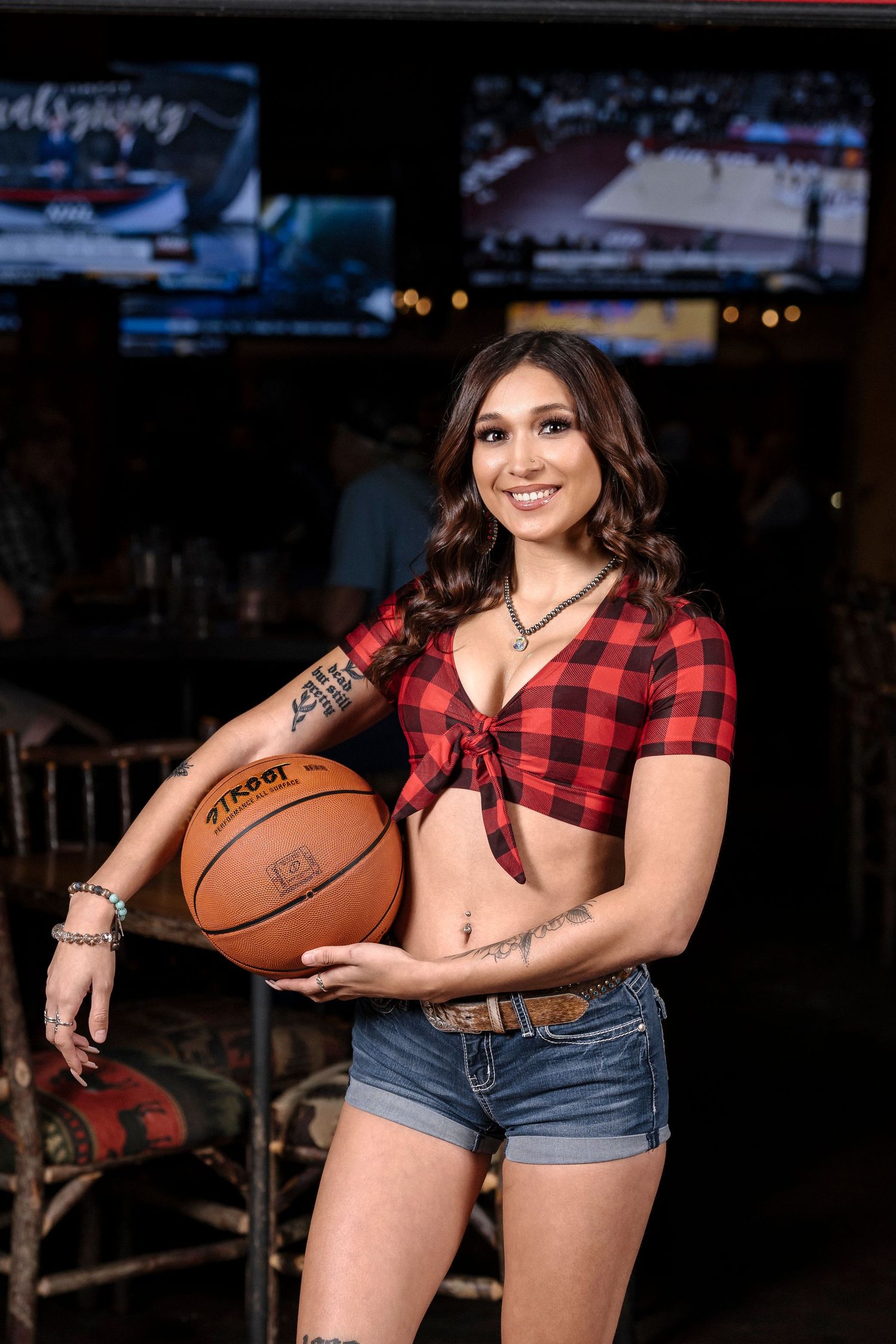 Athletic model in red plaid crop top holds basketball in sports bar setting.