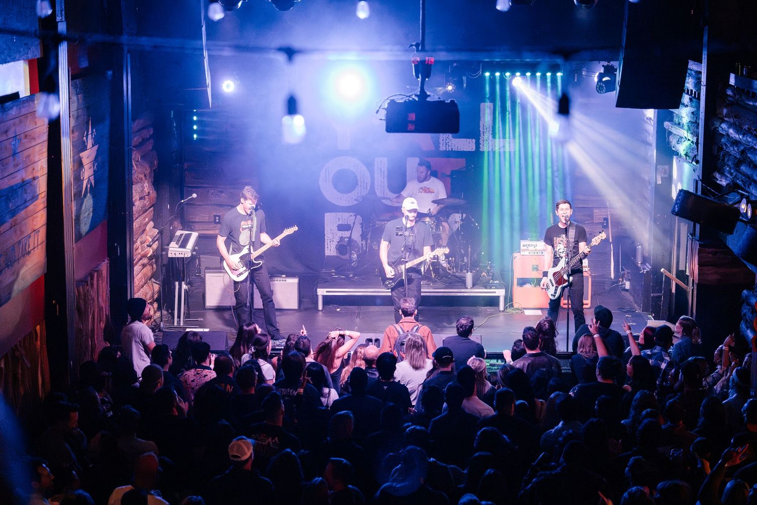 A rock band performs on stage with blue lighting and a packed crowd at a live concert venue.