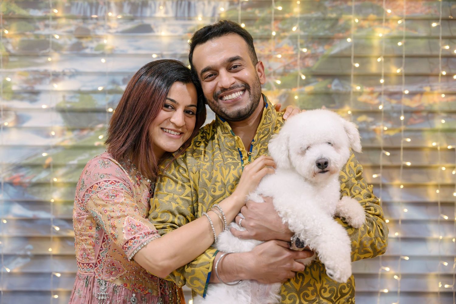 A couple in festive attire holding a white fluffy dog against a decorated background.