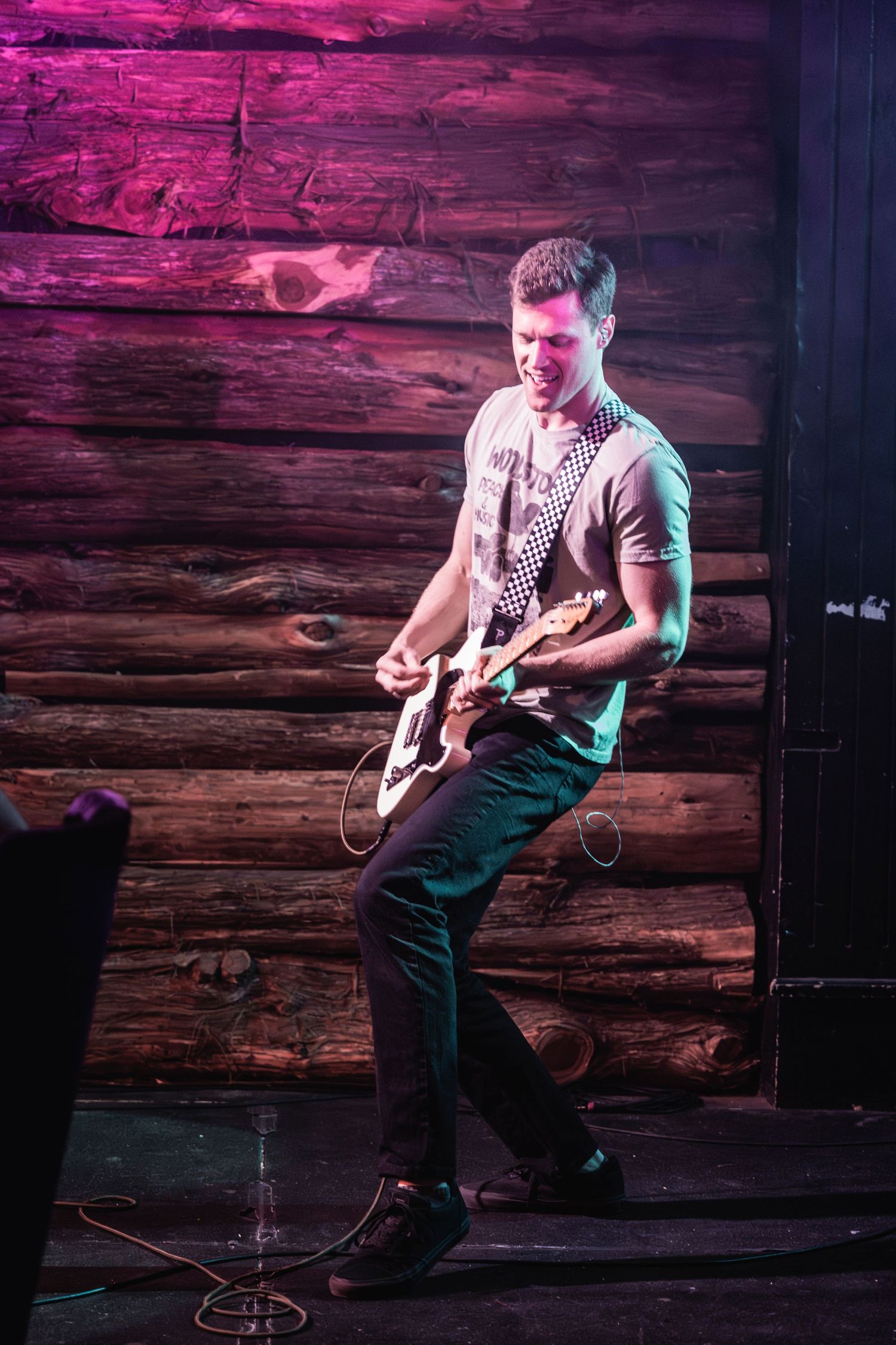 Guitarist performing on wooden stage under dramatic pink and purple lighting.