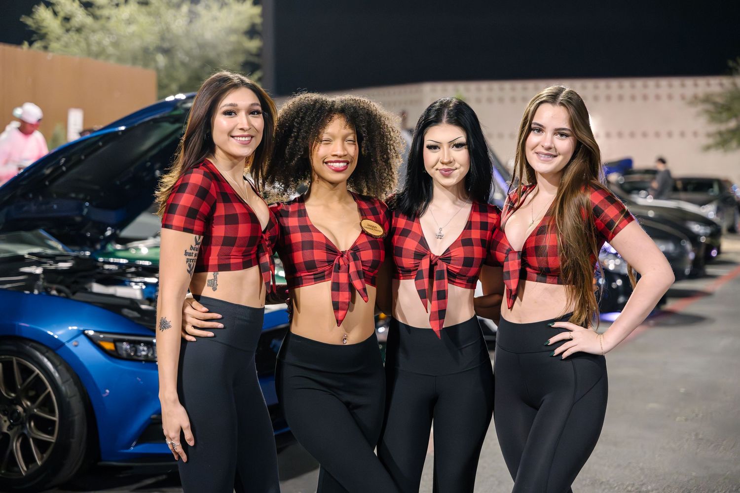Four people wearing matching red plaid crop tops pose together in front of blue car.