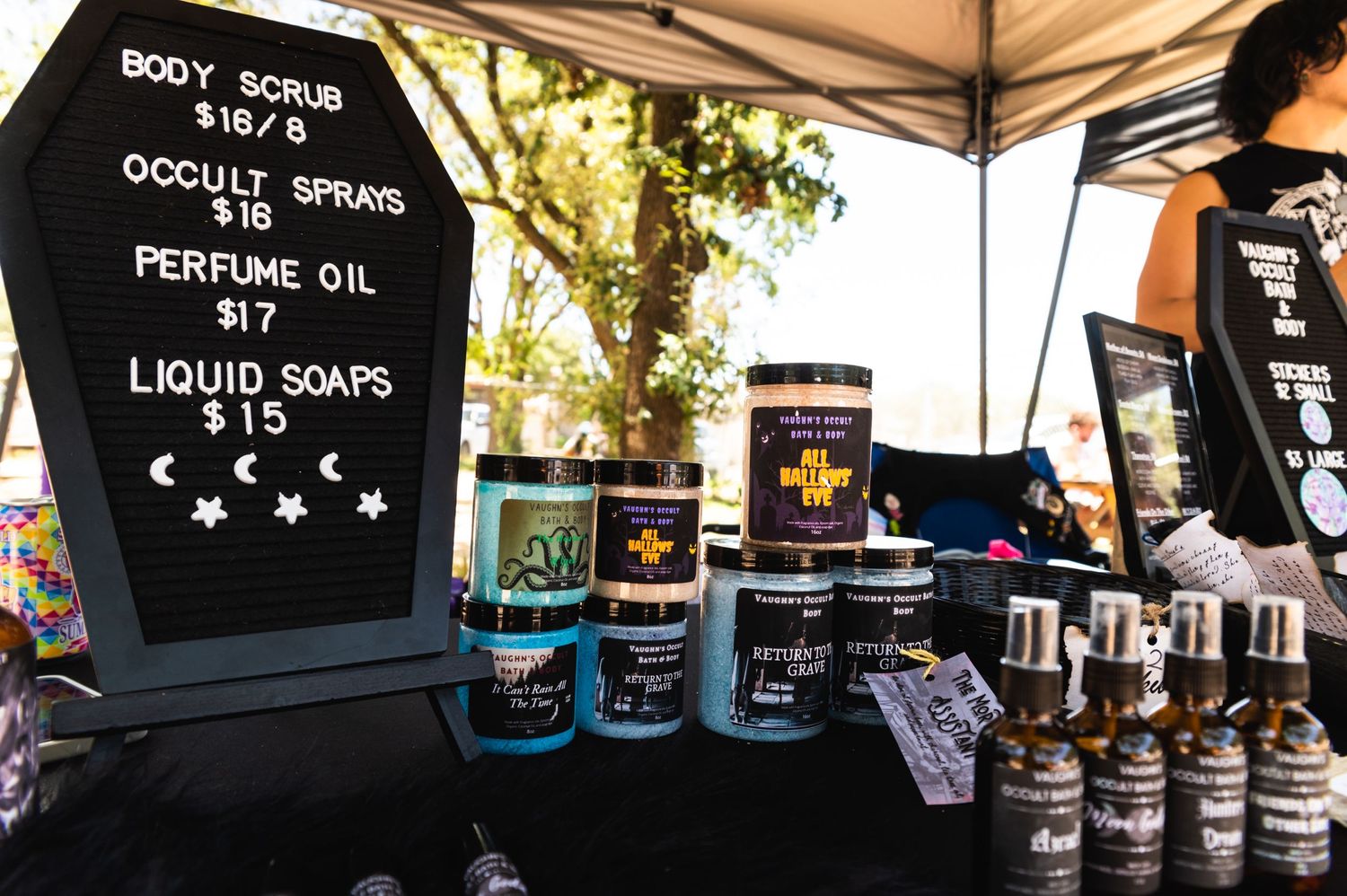 Vendor booth selling body scrubs, perfume oils and liquid soaps with price list display.