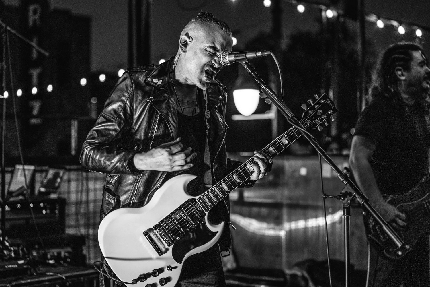 Performer plays white electric guitar during nighttime concert with string lights in background.