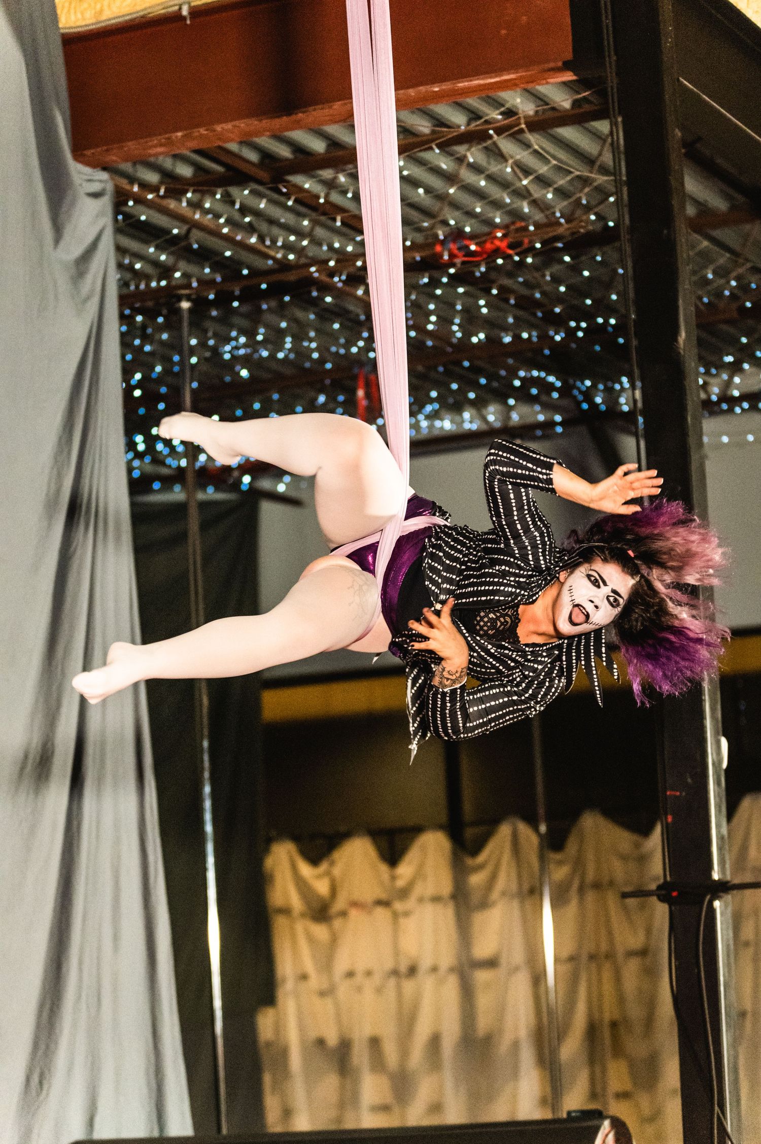Aerial performer in striped outfit performs on pink silk fabric with sparkly lights in background.