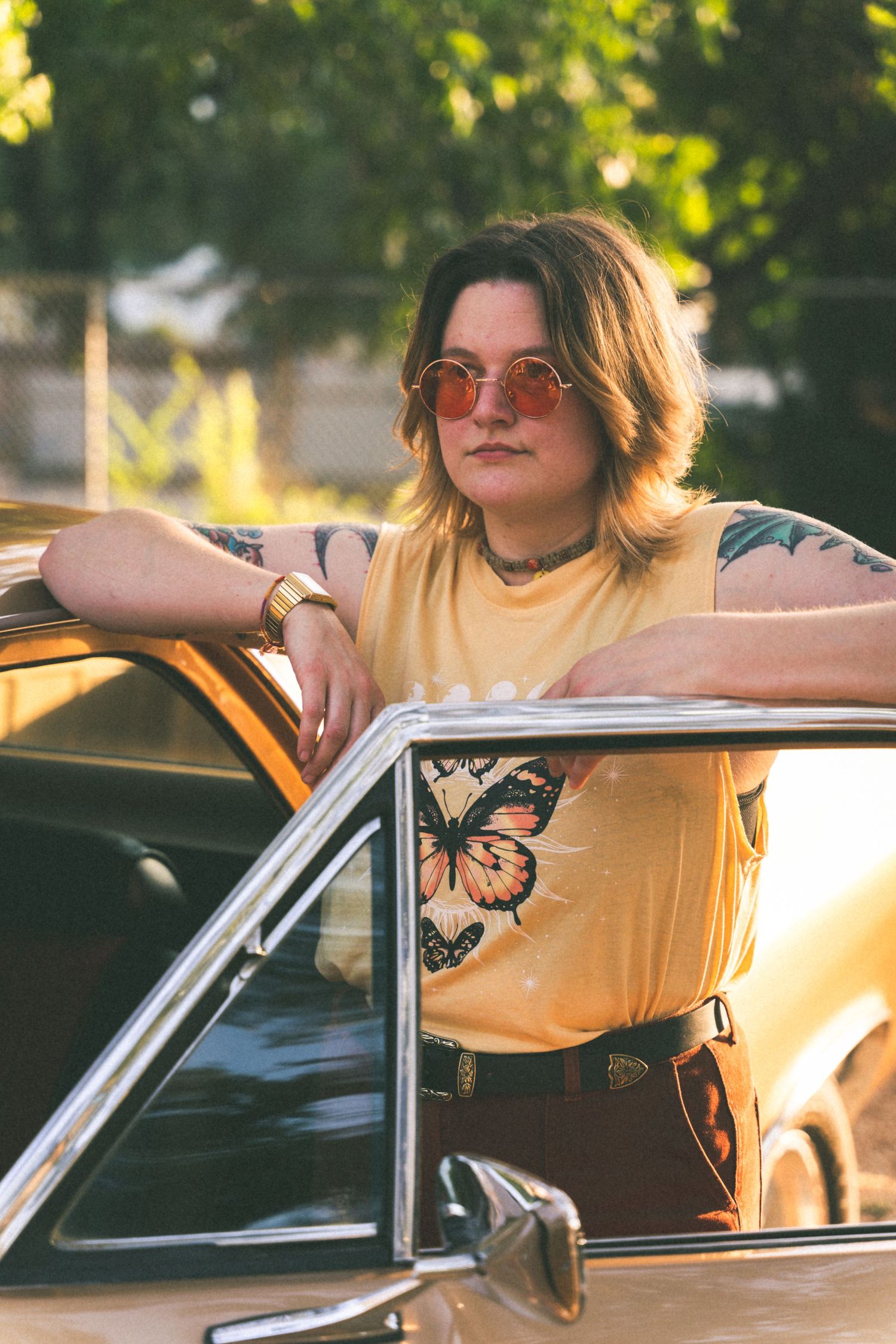 Young woman in yellow t-shirt leaning against vintage car door in warm sunlight.