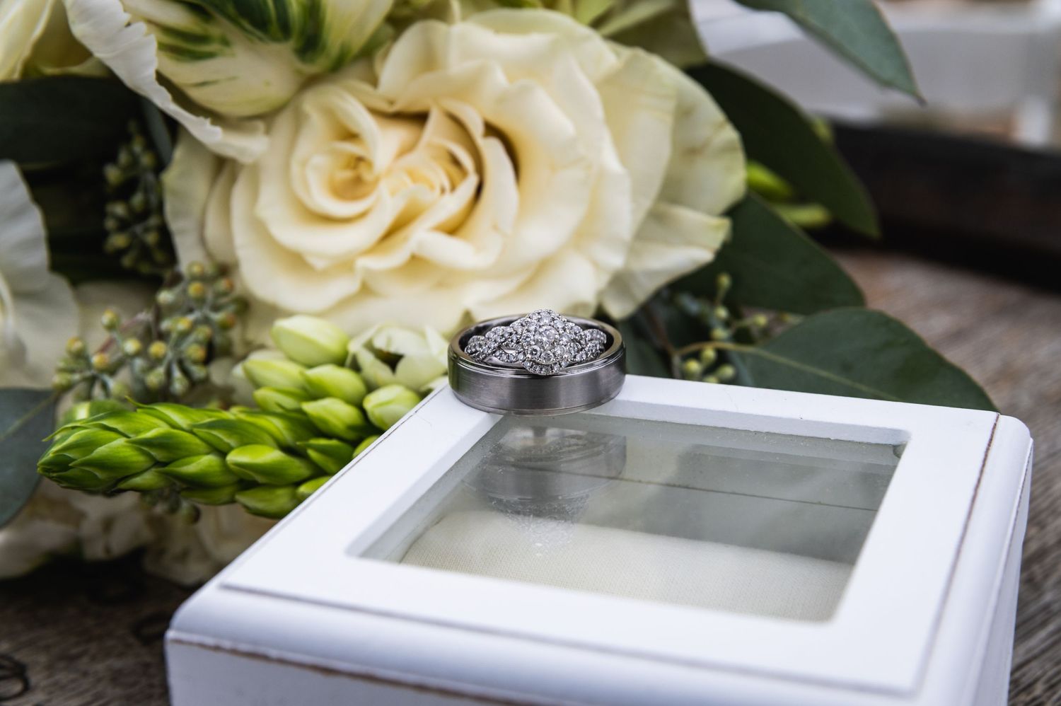 Wedding rings displayed with white roses and greenery on a white display box.