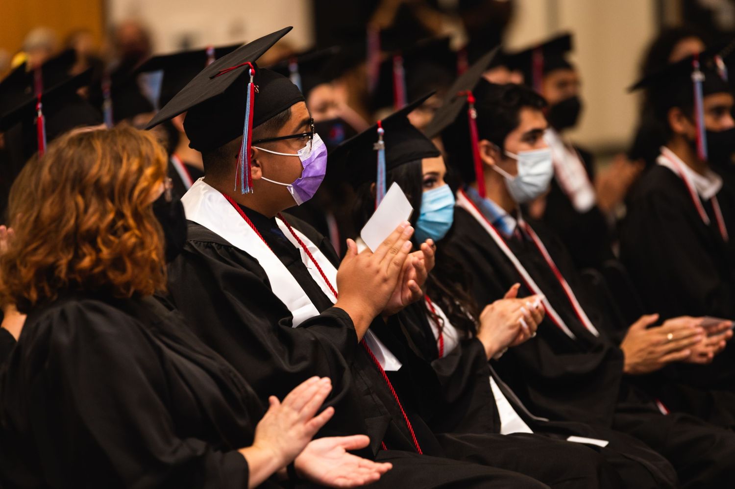 Graduates in black gowns and masks applauding during commencement ceremony.