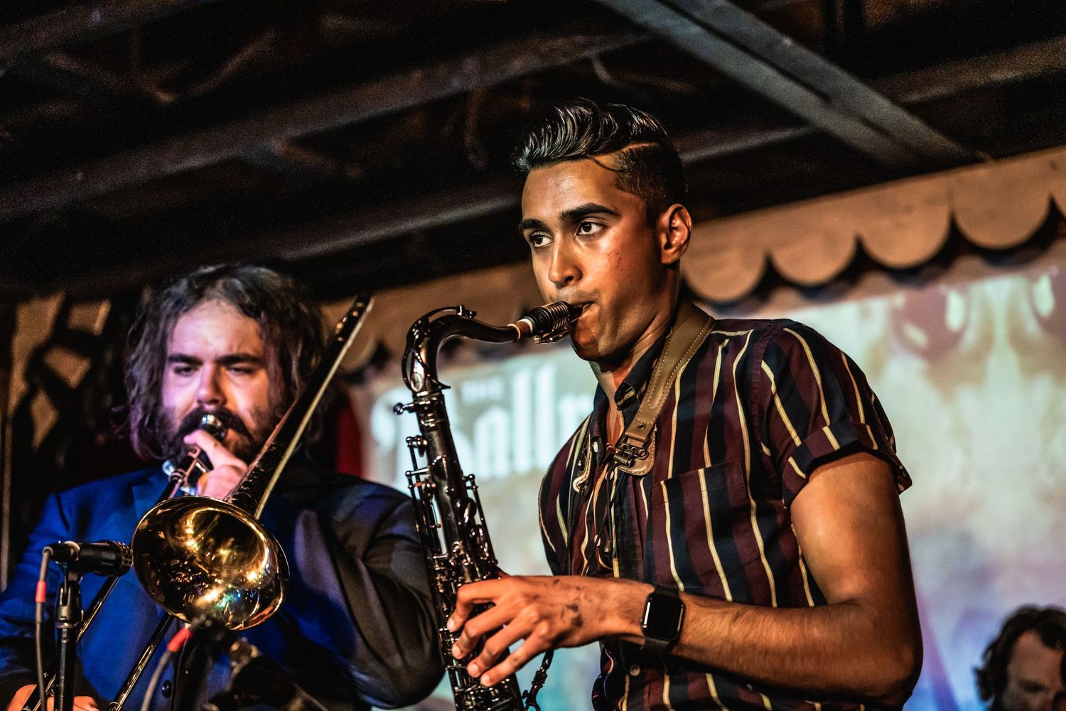 Two musicians perform together, one playing trombone and another on saxophone during a live show.
