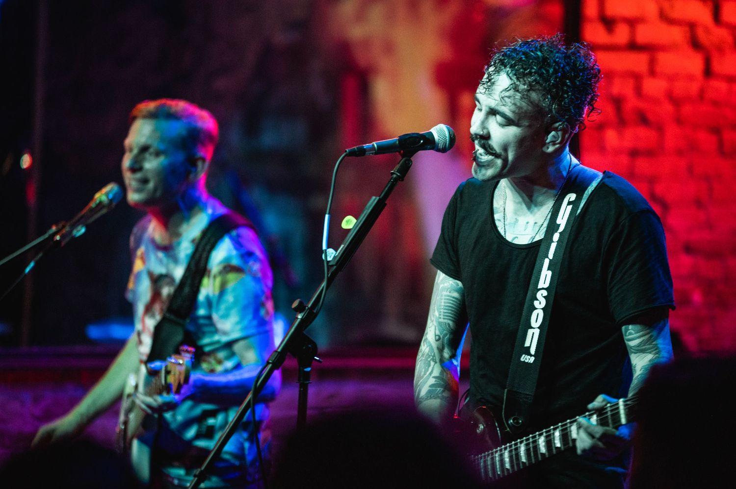 Musicians perform on stage with guitars against a red brick wall backdrop under colorful lighting.