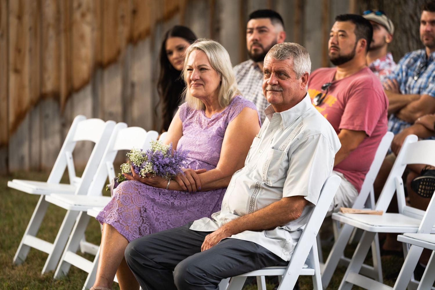 Wedding guests seated in white chairs at outdoor ceremony with wooden barn backdrop.