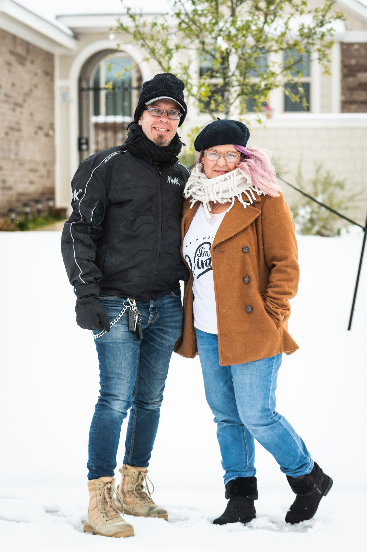 Two people in winter clothing stand smiling together in front of a snowy building.