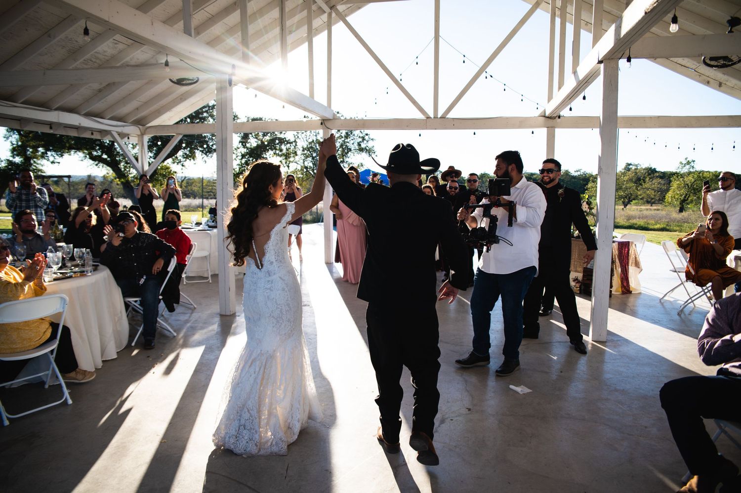 Wedding celebration with guests dancing and cheering under a rustic pavilion at sunset.