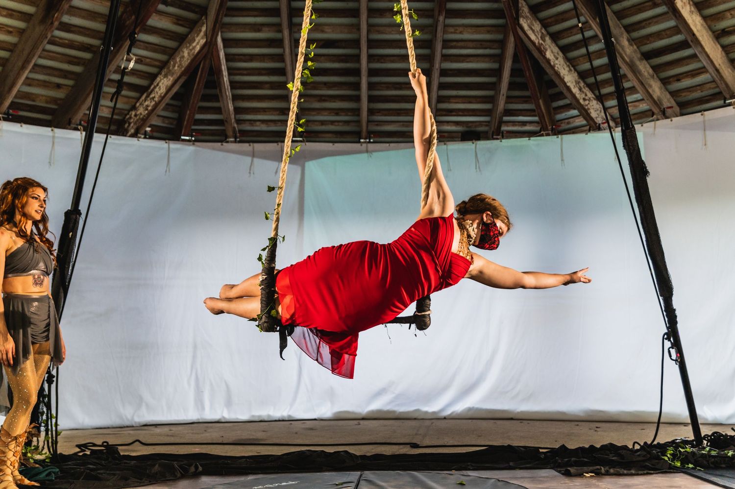Aerial performer in red attire demonstrates horizontal pose on suspended straps against white backdrop.