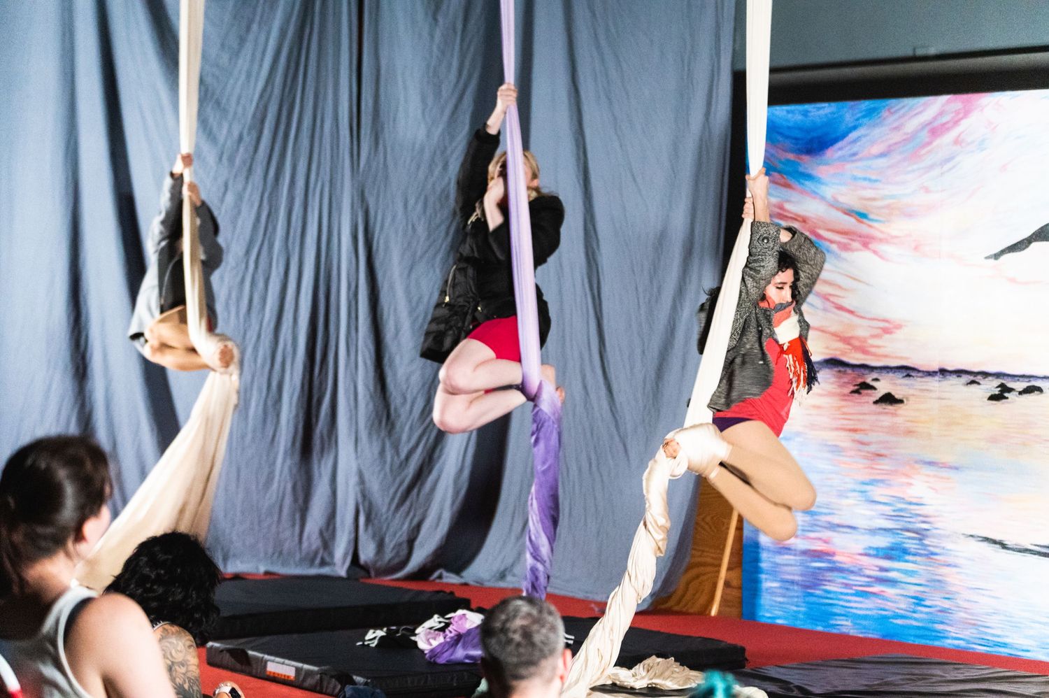 Multiple performers practice aerial silk routines in a studio with blue artwork in background.