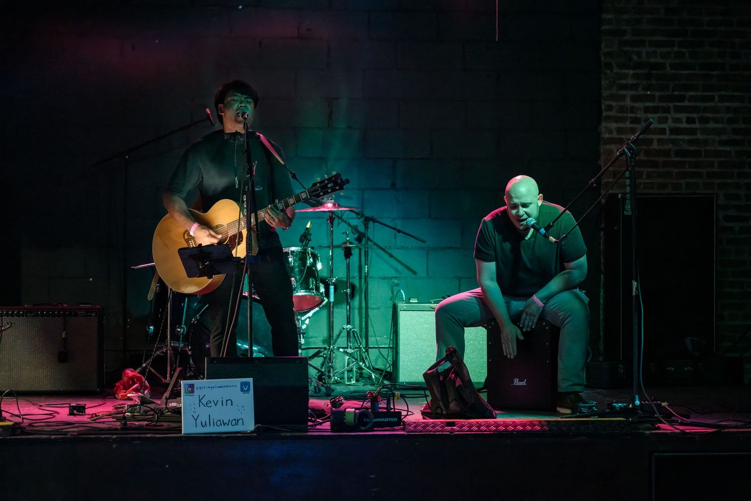 Musical duo performs acoustic set with guitar and percussion under dramatic blue-green lighting.