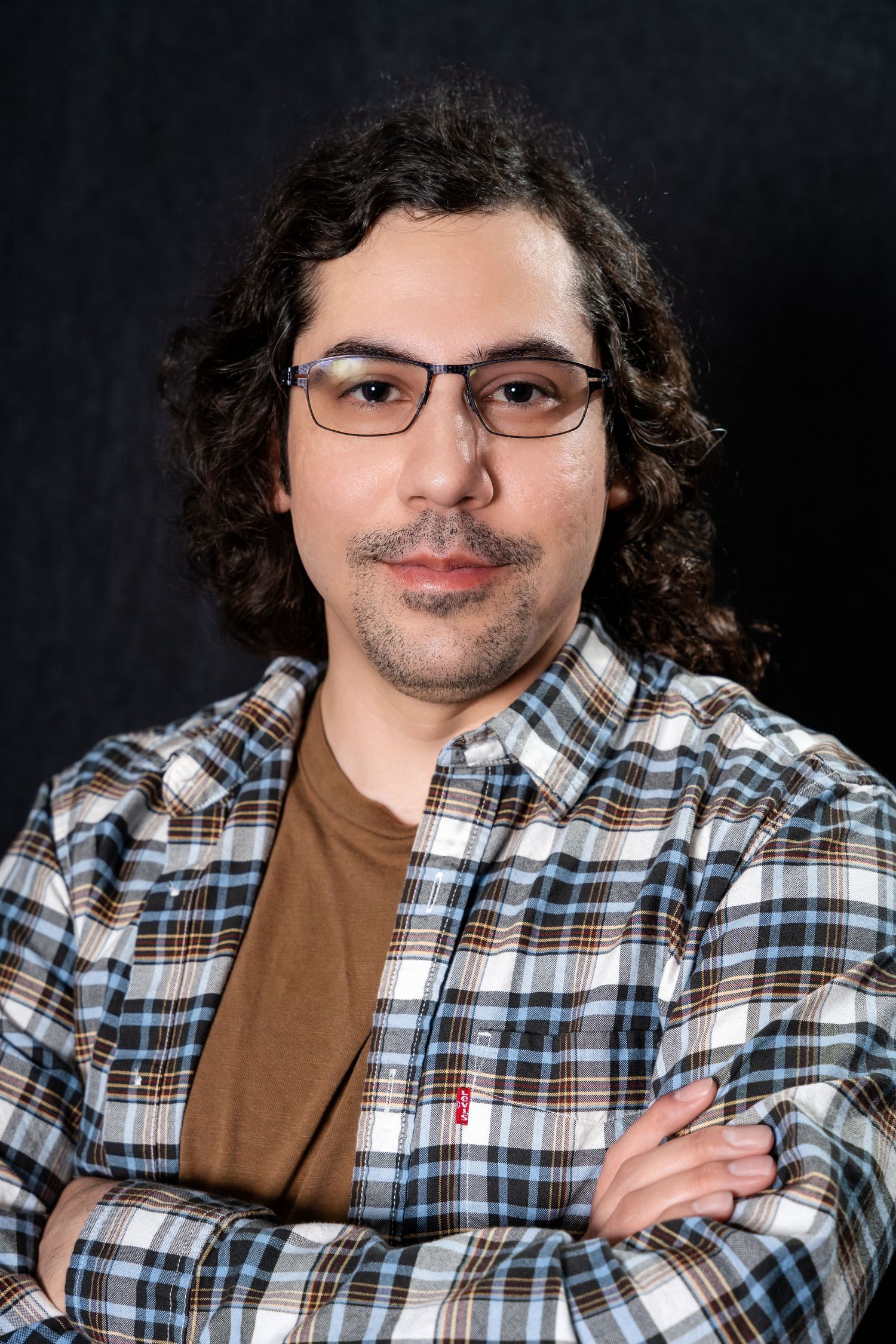 Studio portrait of a person in plaid shirt and glasses with curly hair against black backdrop.