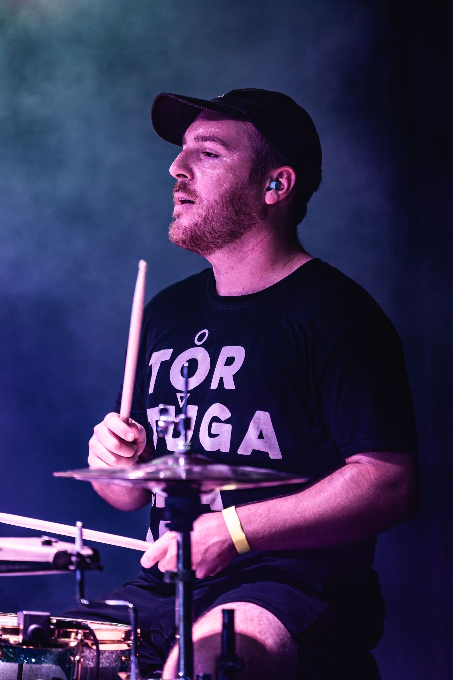 Drummer performs on stage wearing a black t-shirt under atmospheric blue lighting.