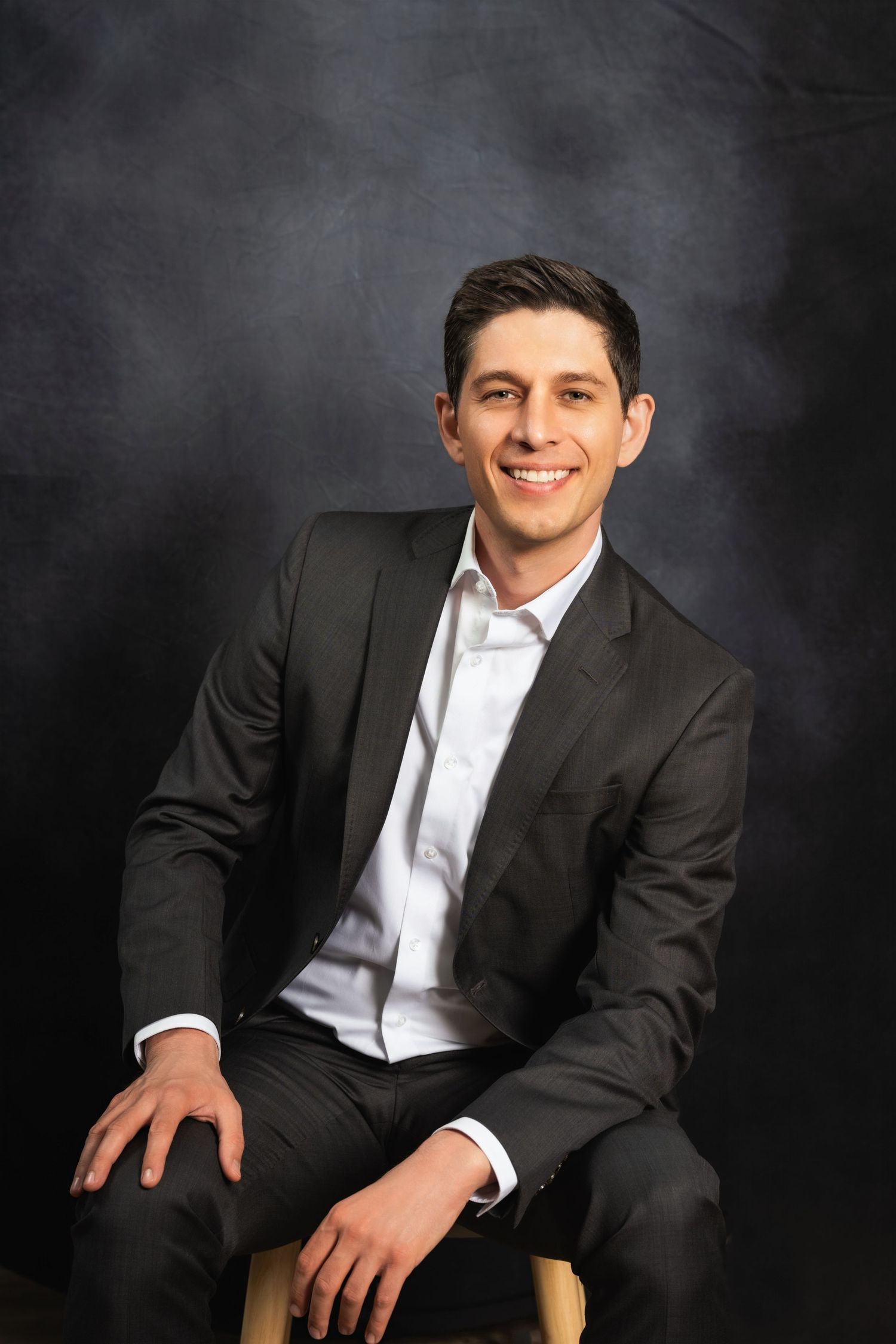 Corporate headshot of person in business attire smiling against dark background.