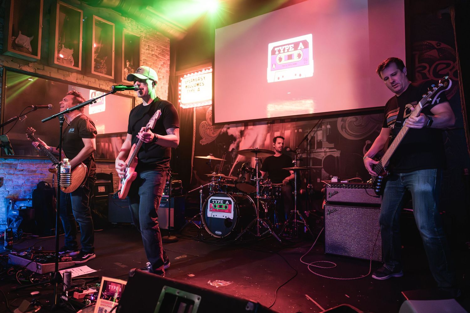 Band performs on dimly lit stage with projection screen and musical equipment visible.