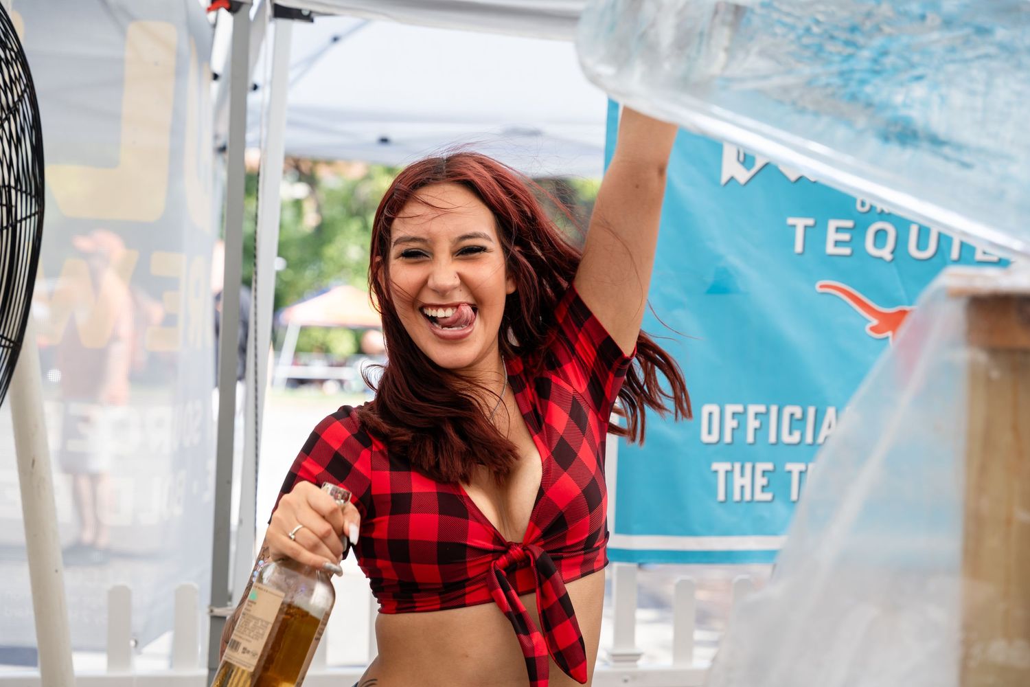 Event staff member in plaid top holding bottle at outdoor promotional tent.