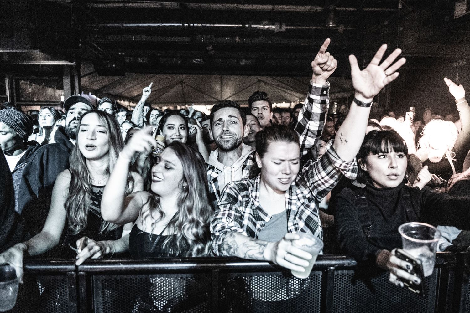 An enthusiastic crowd at a concert raising their hands and cheering in black and white.