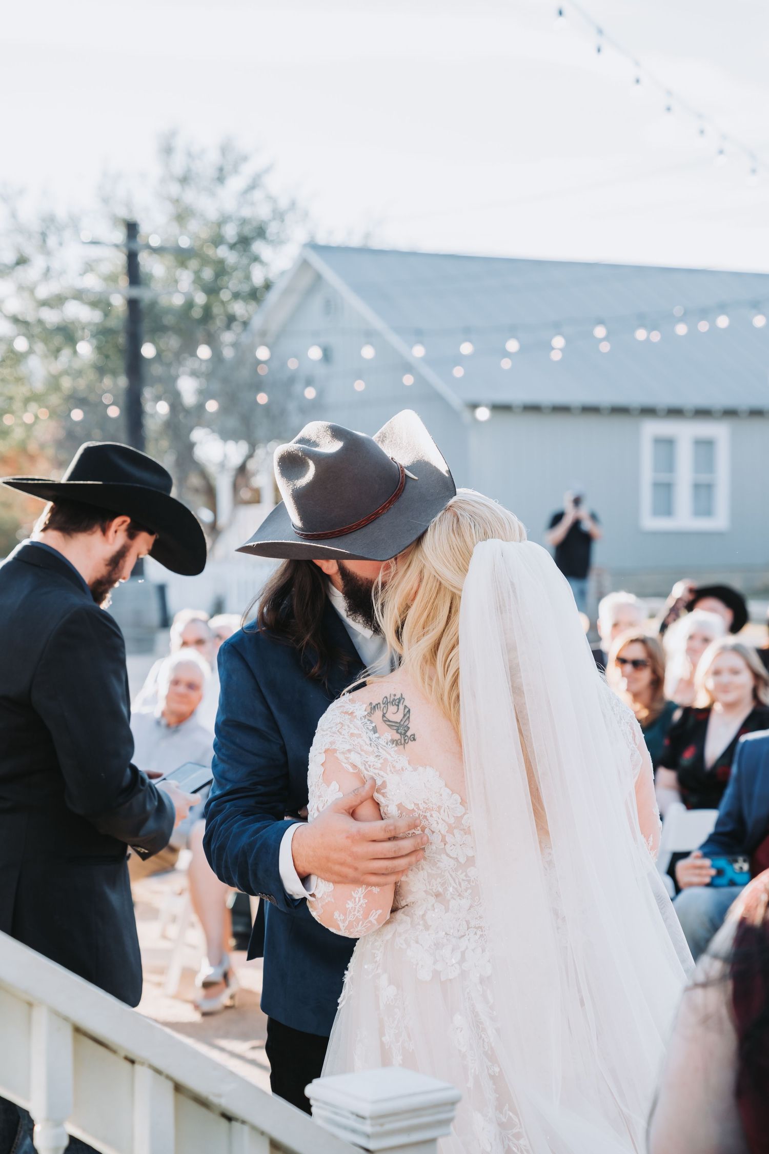 An outdoor wedding ceremony with string lights and guests seated on white chairs.