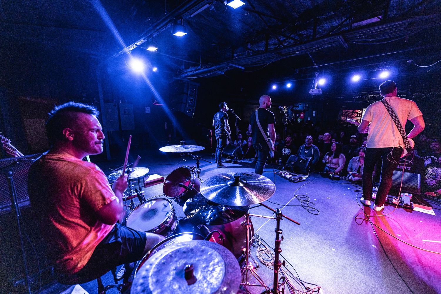 Drummer plays on stage with blue lighting while band performs in background.