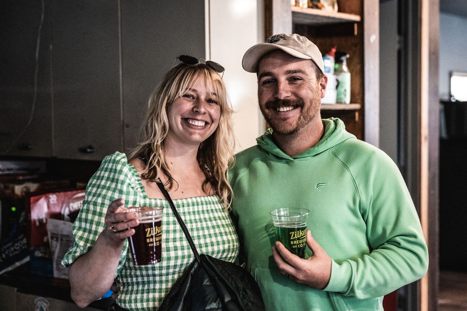 Two people in casual attire share drinks and smiles in kitchen setting.