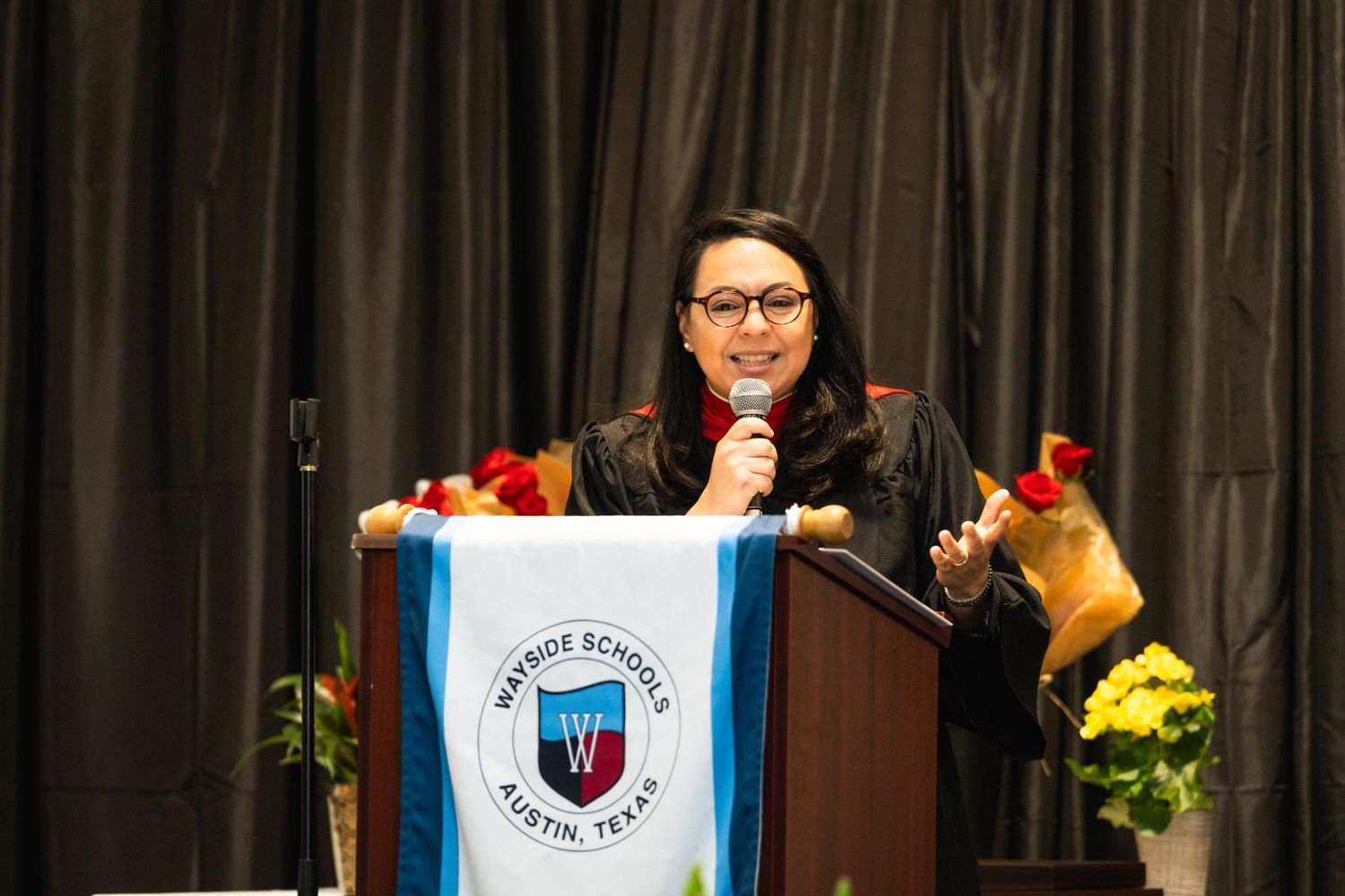 Speaker at graduation podium with blue and white banner and decorative flowers.