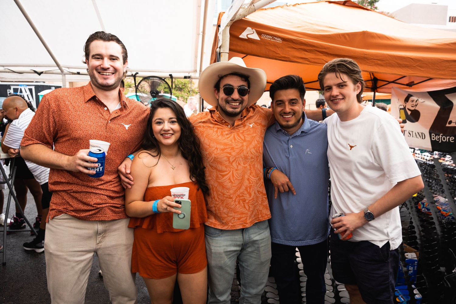 Group in orange and casual attire posing together under a white tent at an outdoor gathering.