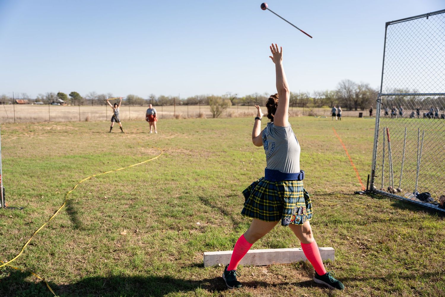 Athlete in kilt and red socks throws hammer at Highland Games competition on green field outdoors.