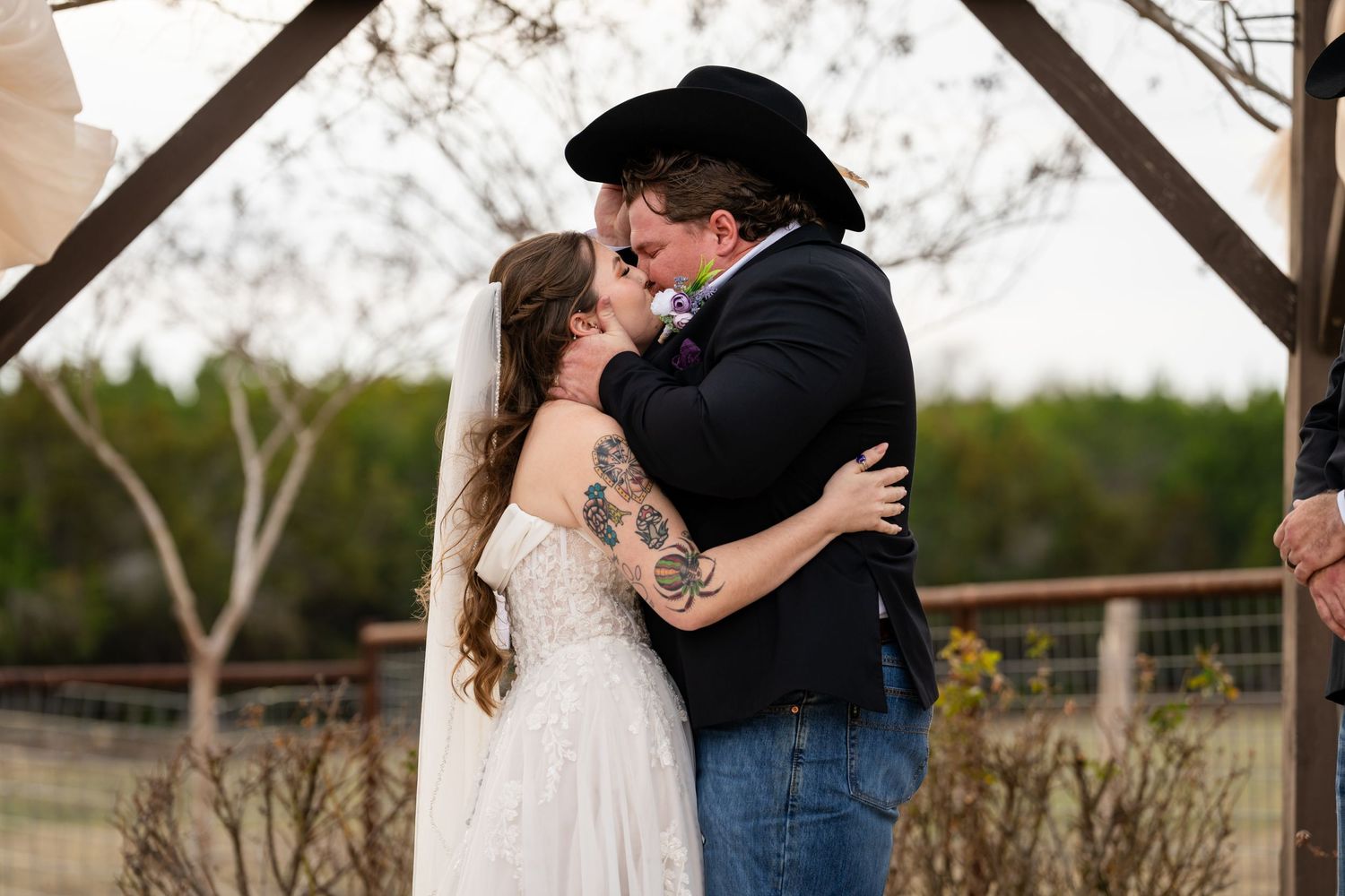 Bride and groom share first kiss at outdoor rustic wedding ceremony, groom wearing black cowboy hat and jeans.