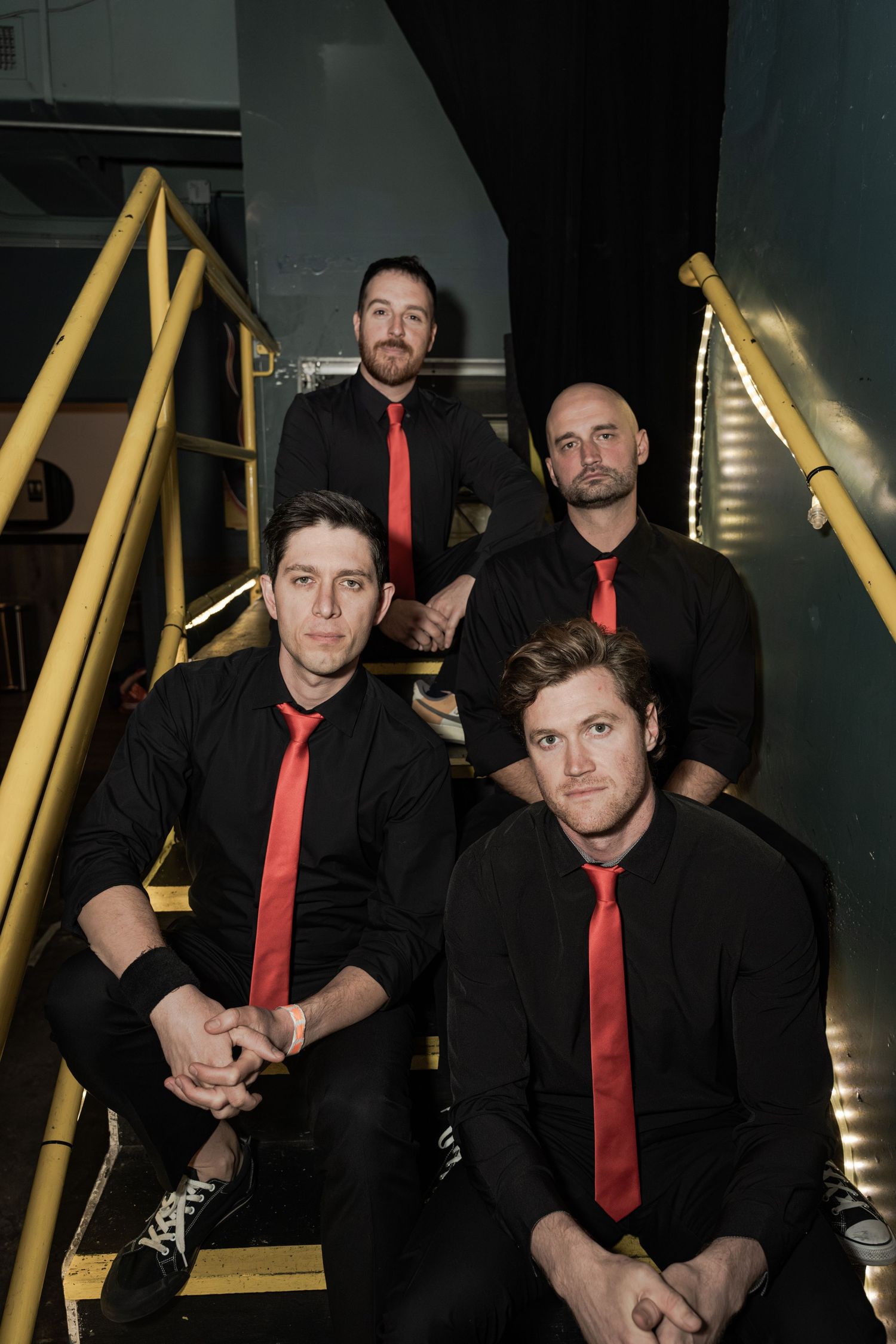 Four men in matching black shirts and red ties pose on yellow stairs for a professional band photo.
