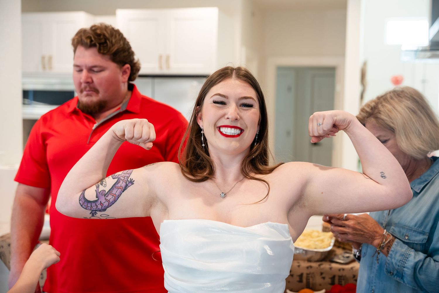 Woman in white strapless dress flexing bicep tattoo, smiling, with two people in background at indoor gathering.