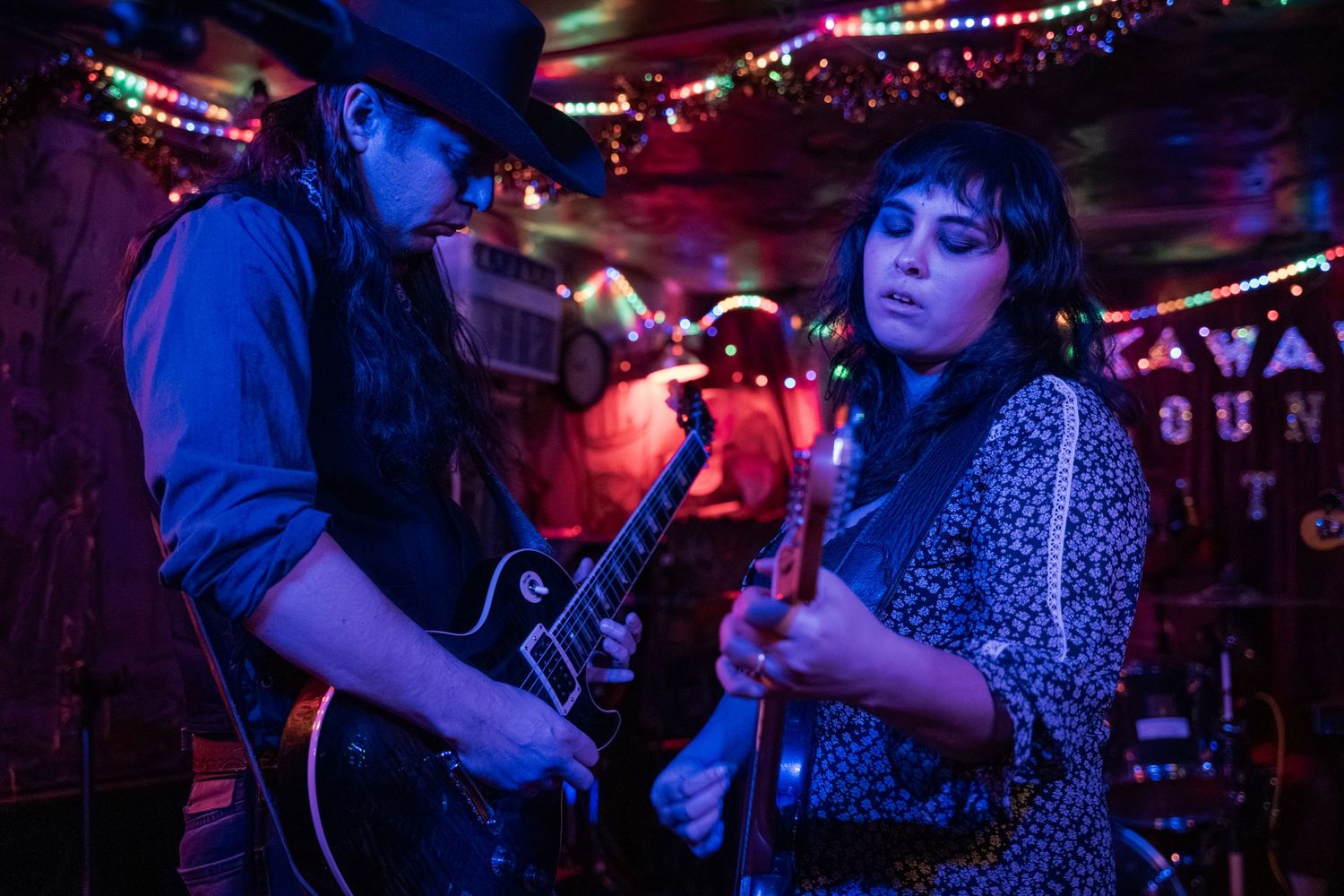 Musicians perform on stage at a dimly lit bar venue with colorful string lights creating an intimate atmosphere.
