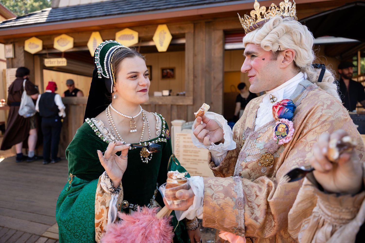 Two people in ornate Renaissance period costumes interact while enjoying refreshments at an outdoor festival.