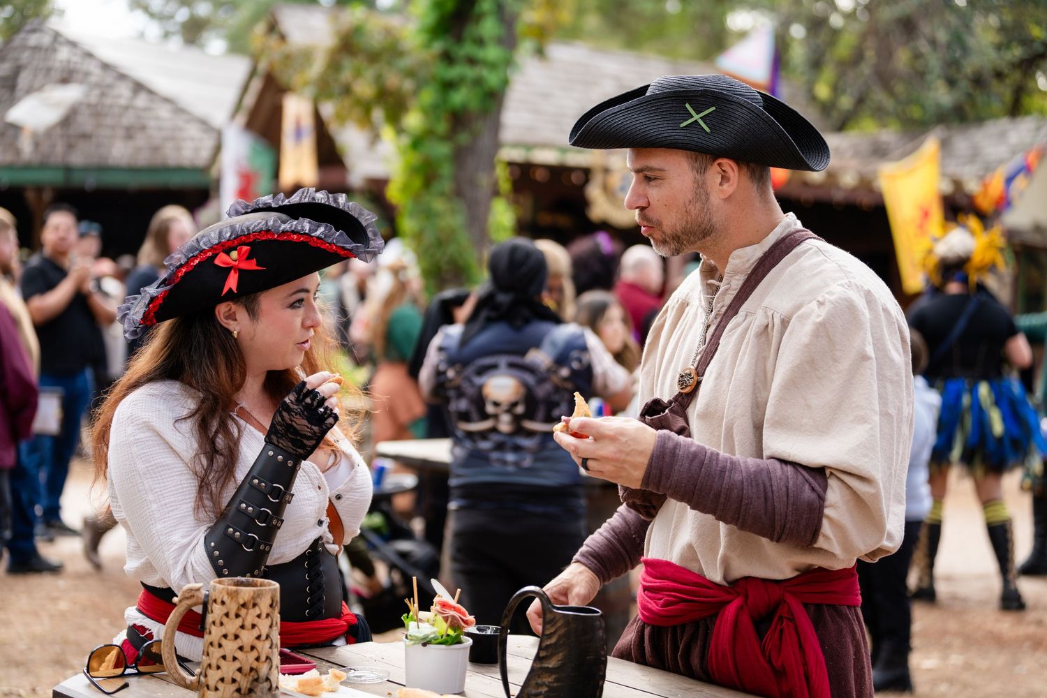 Two people in Renaissance festival costumes interact at an outdoor fair with other attendees visible in background.