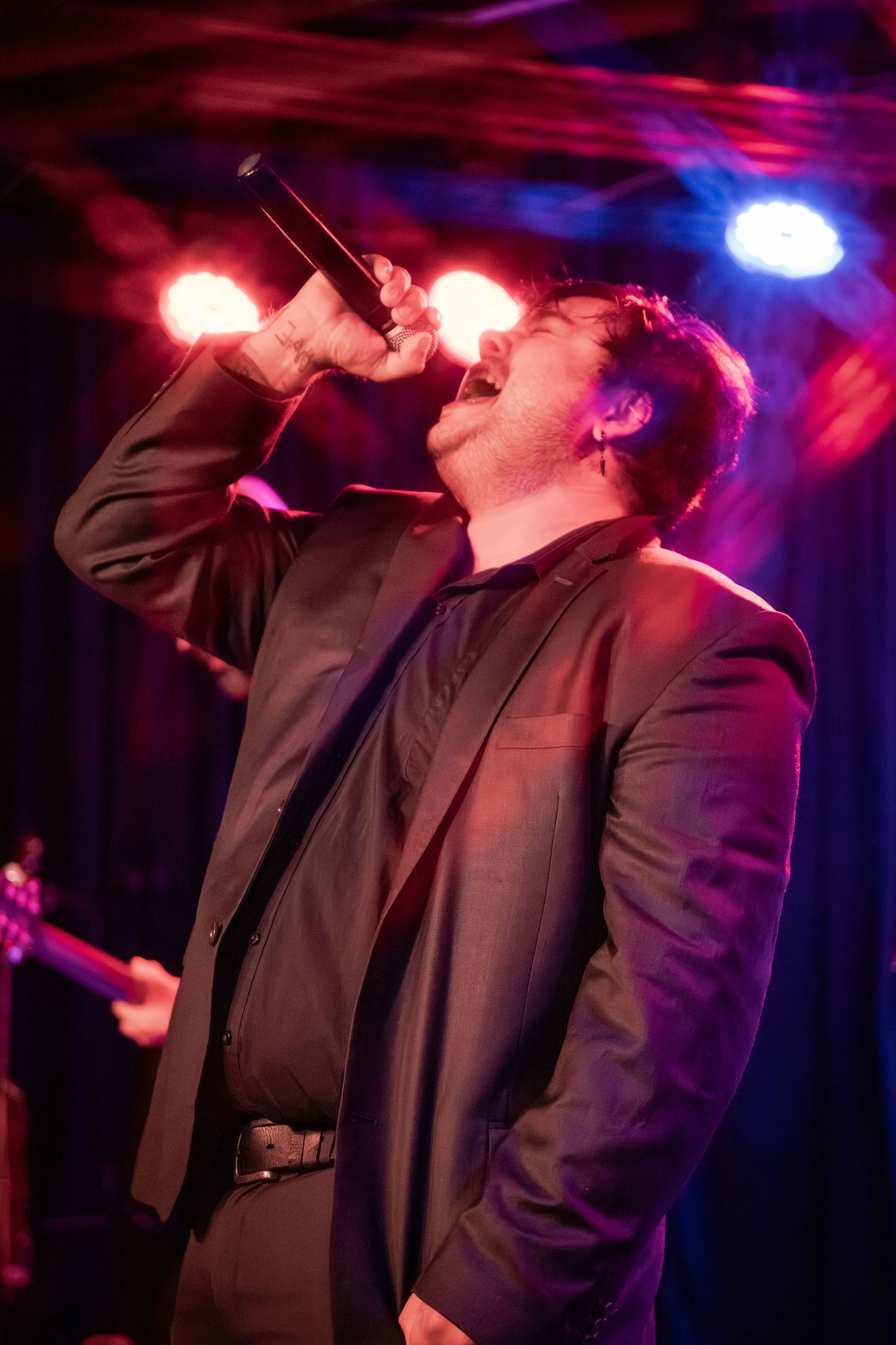 A performer in a dark suit sings passionately on stage under dramatic red and blue lighting at a nightclub venue.