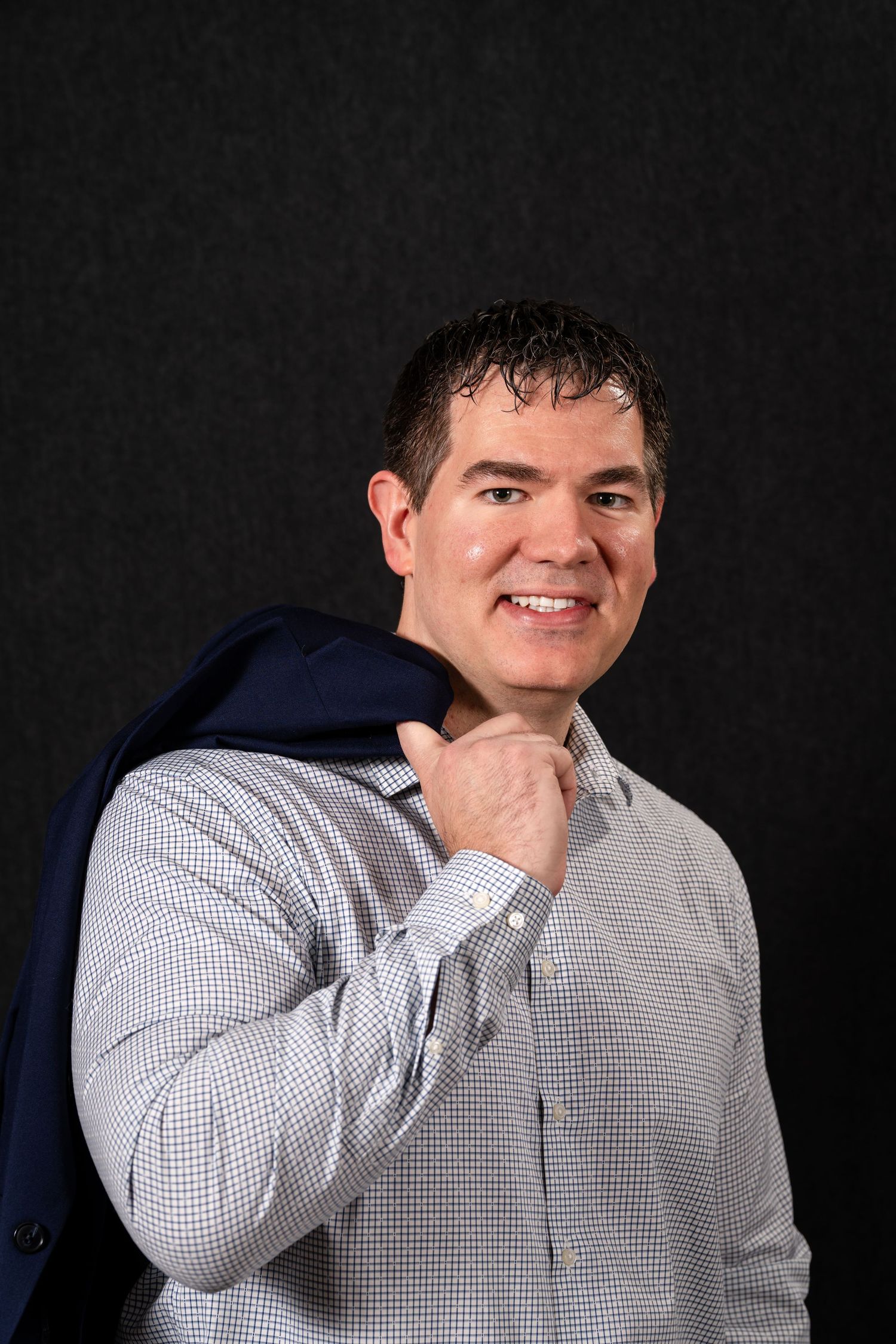 A person in a checkered button-down shirt poses against a dark background while covering their mouth.