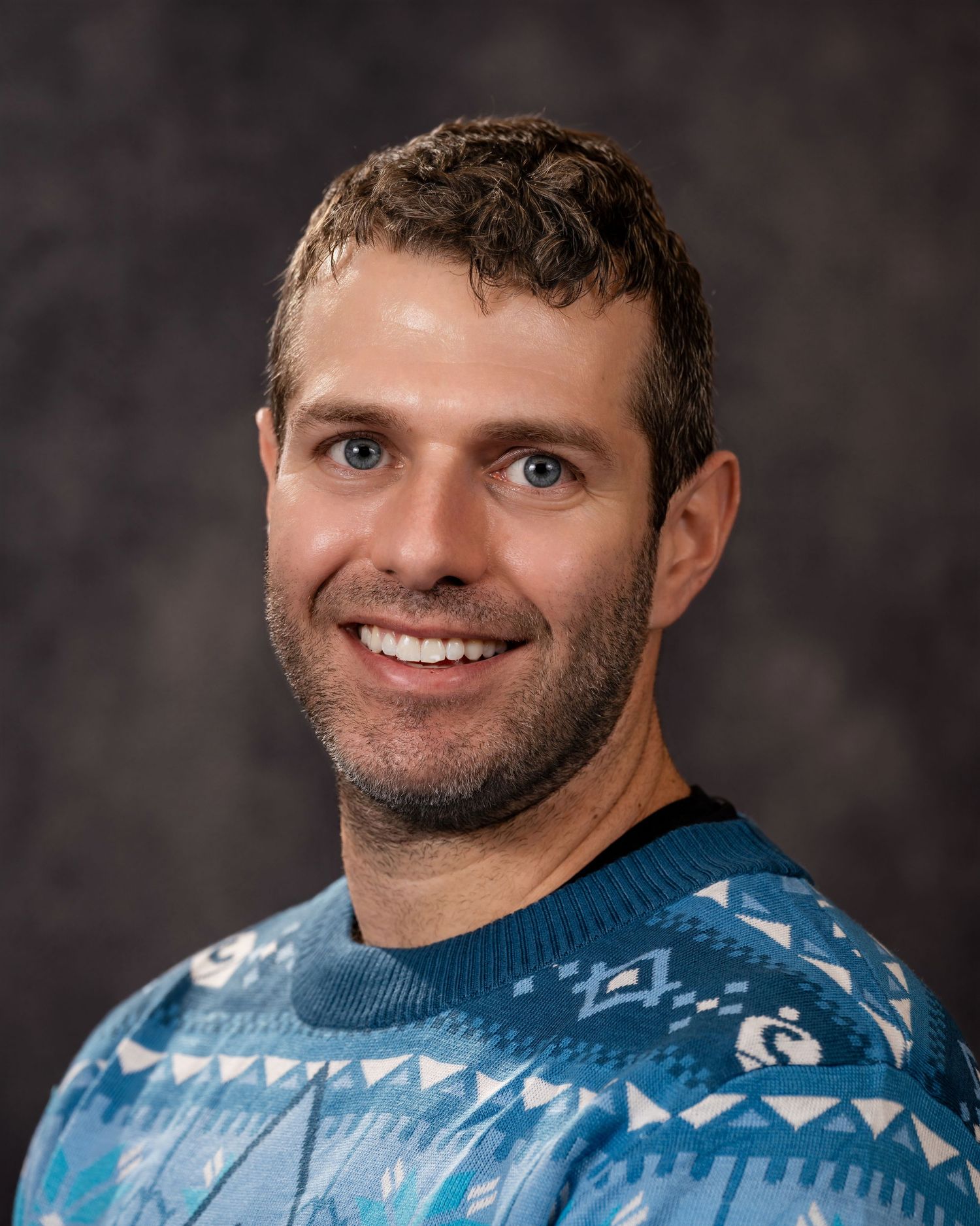 A person in a light blue patterned athletic shirt smiles warmly against a dark background.